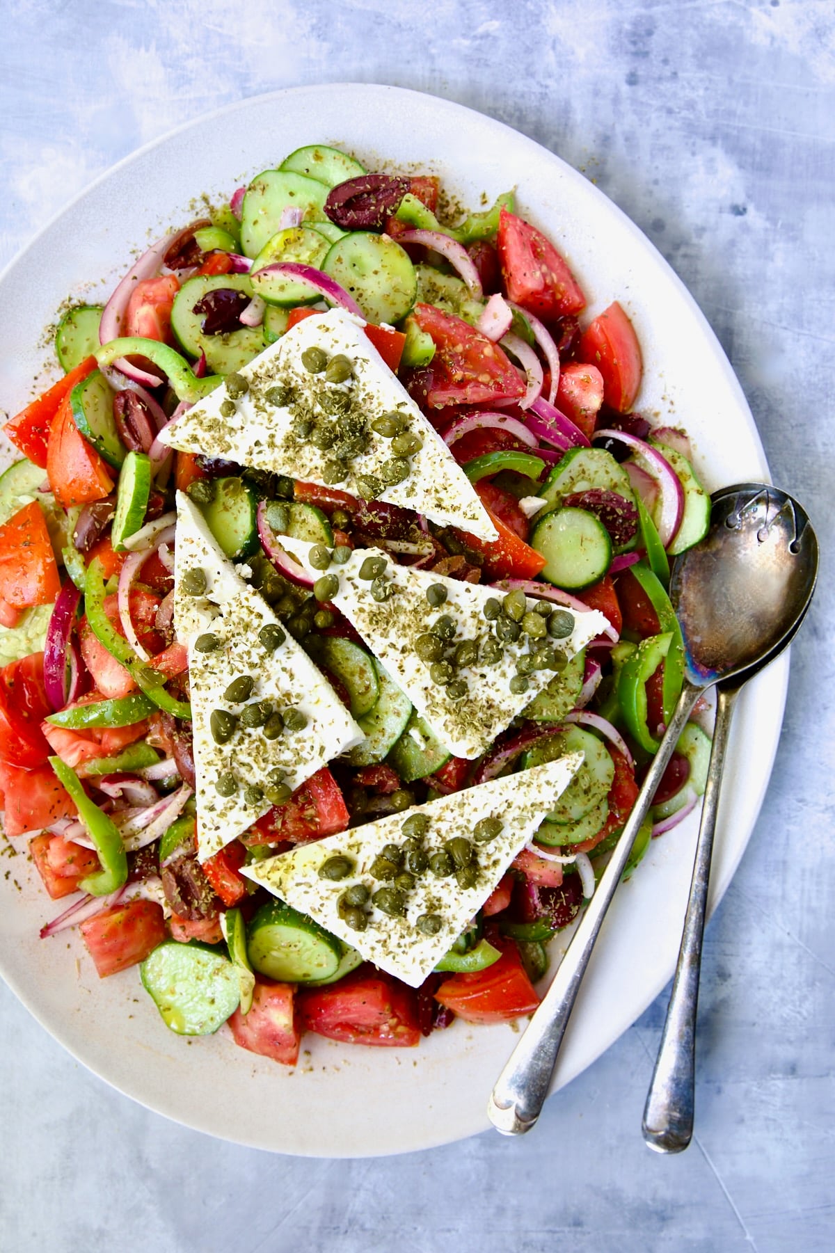 Classic Greek Salad with four triangles of feta cheese on white oval platter with serving utensils.