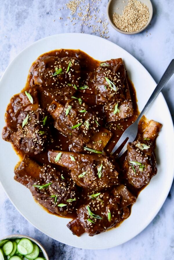 Korean Braised Beef Short Ribs on oval white platter with meat fork sprinkled with sesame seeds and thinly sliced scallions.