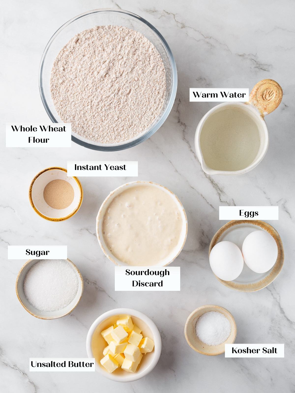 Ingredients for whole wheat sourdough rolls arranged on countertop including flour, butter, and eggs.