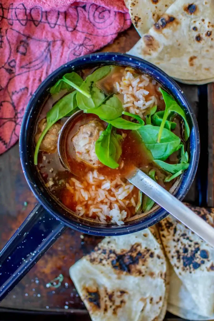 A small blue pot filled with meatball soup with rice and arugula leaves with a ladle in it.