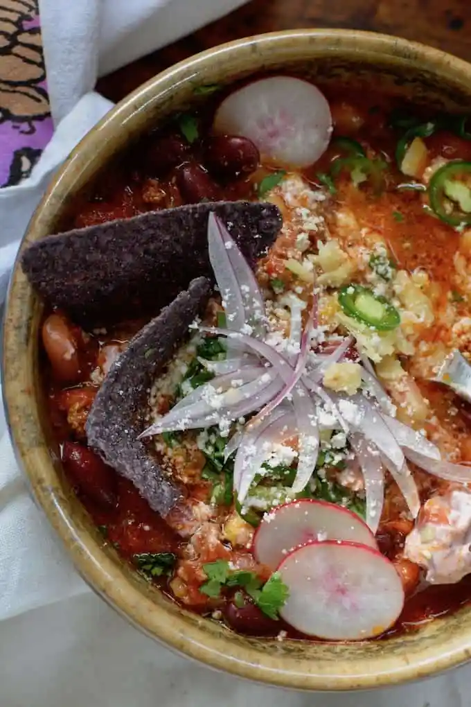 A bowl of chili topped with thinly sliced red onions and blue tortilla chips.