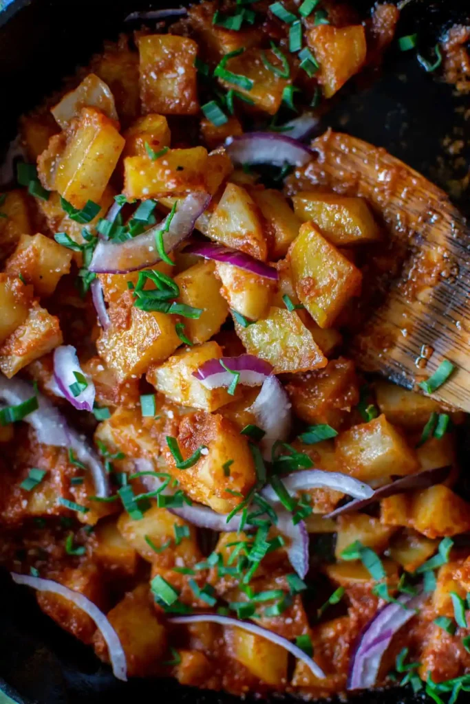 A close up on a pan of potatoes covered in red sauce being stirred with a wood spoon.