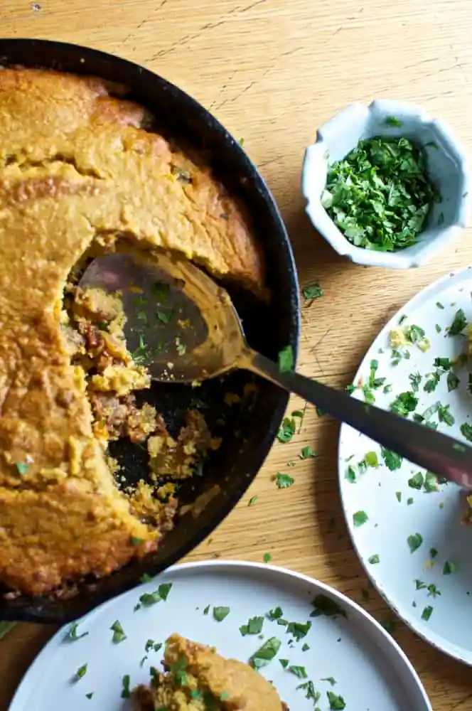 A casserole topped with a sweet potato corn bread crust with a scoop taken out of it and a spoon in the pan.