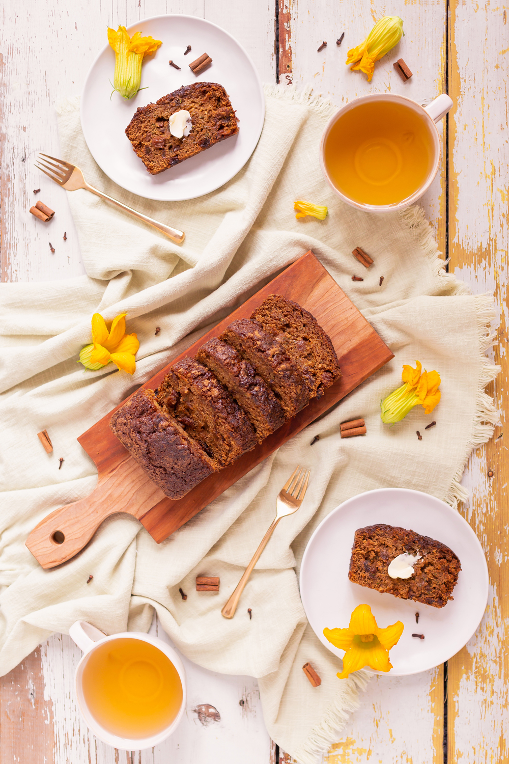 Cinnamon Raisin Zucchini Bread {Dairy Free, Gluten Free, Refined Sugar Free} | Sliced Zucchini Bread on a wooden serving board, served on 2 cream stoneware plates with gold forks, squash blossoms and golden tea | Moist Zucchini Bread Recipe | Easy Cinnamon Raisin Loaf | Best Baking Recipes for Overgrown Garden Zucchini | Zucchini Bread Cake | Healthy Cinnamon Zucchini Bread | What to do with large zucchini | Zucchini Bread with Crispy Top | Calgary Plant Based Food Blogger // JustineCelina.com