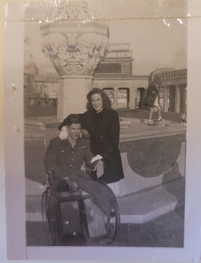 Black and white photo shows man wearing uniform sitting in a wheelchair and a woman in dark coat standing beside him with arm around shoulder. In the background is what appears to be a decorative fountain.
