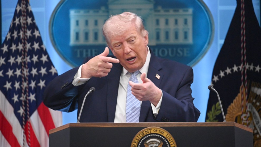 Trump mimics shooting a rifle while standing at a podium and speaking into a microphone at a press conference. He is wearing a black suit and a light blue tie.