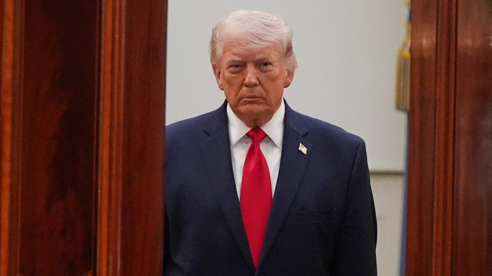 Donald Trump stands by an open doorway with a blank expression. He is wearing a black suit, red tie, and an American flag pin on his left side. The doors are reddish-brown.