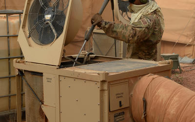 A man in brown camouflage clothing, including baseball cap, gloves and ear protection, uses a long pole to clean the inside of an HVAC unit outside a military tent.