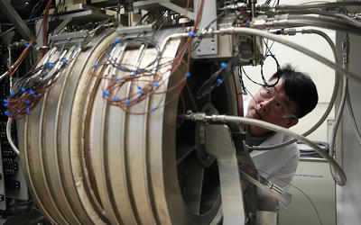 A researcher peers around the edge of large cylindrical piece of equipment in the lab. 