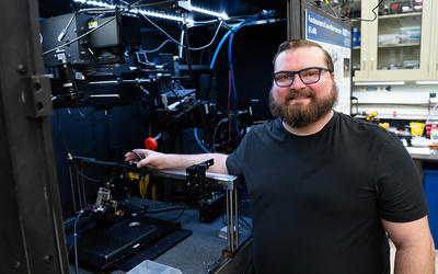 A bearded man poses smiling next to an open cabinet of equipment in a lab. 
