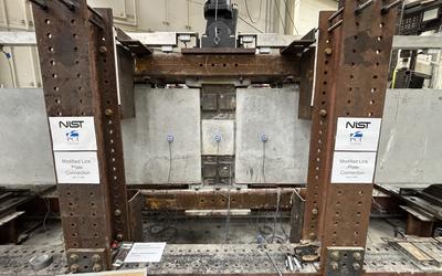 A wide view of a concrete beam assembly inside a large metal frame. A hydraulic arm is part of the metal frame, and it’s posed to press down at a section in the middle where the concrete pieces connect.