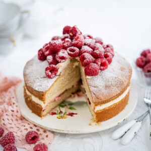 A Victoria sponge air fryer cake topped with fresh raspberries and powdered sugar, with a slice removed to reveal cream and jam filling. The cake sits on a floral plate, with scattered raspberries and decorative forks beside it.