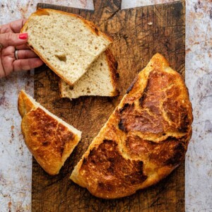 A round loaf of crusty Quick Sourdough Bread With Yeast sits on a wooden cutting board. Two slices are cut from the loaf; a hand with red nail polish holds one slice. The background is a white, textured surface.