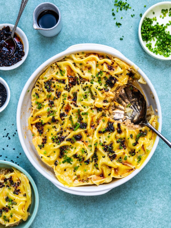 A baked Dumpling lasagne in a round white dish, with wavy wonton wrappers, melted cheese, pork filling, and sprinkled herbs. Small bowls with sauces and chopped chives are placed nearby on a light blue surface.