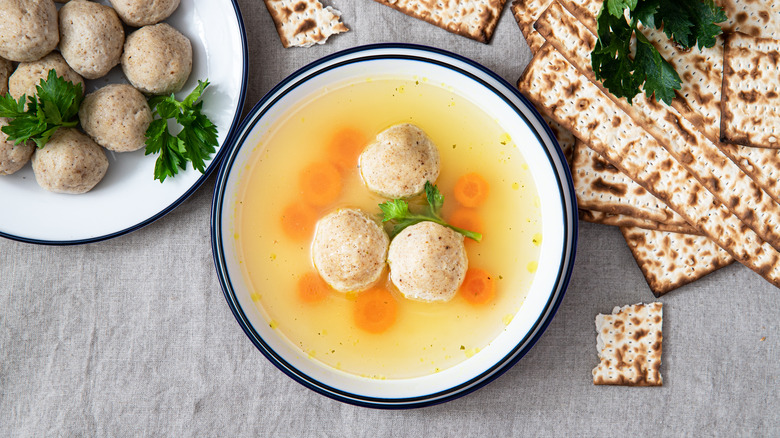 Matzo ball soup in a bowl on a table with matzah and additional dumplings on a plate