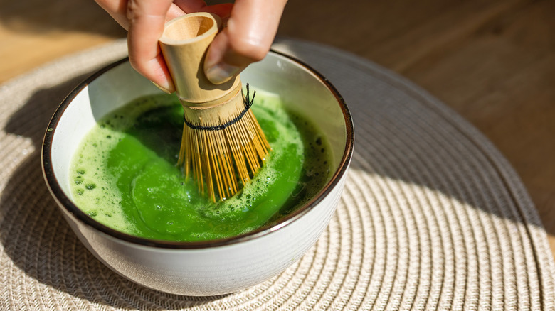 Hand holding bamboo chasen whisk to mix matcha green tea inside white bowl