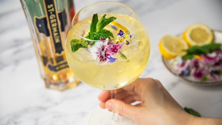 Hand holding springtime Hugo Spritz with mint, edible flowers and lemon slice