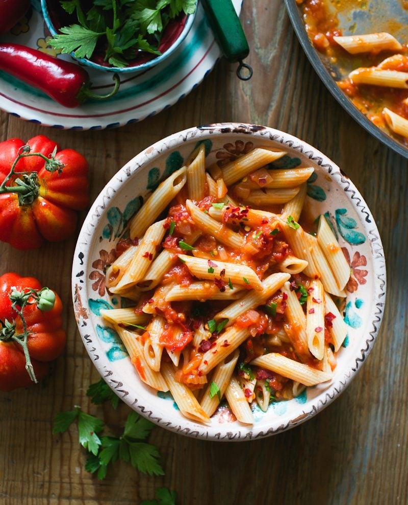 Arrabbiata sauce with penne pasta in a bowl with fresh tomatoes and parsley in the background.
