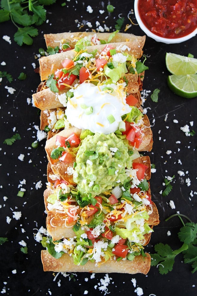 These easy oven Baked Chicken Taquitos are stuffed with chicken and cheese and make a great appetizer or meal. They can be made in advance and are perfect for parties! Serve with guacamole, sour cream, and salsa. Baked Chicken Taquitos with cheese, lettuce, diced tomatoes, cilantro, sour cream, and guacamole on sheet pan.